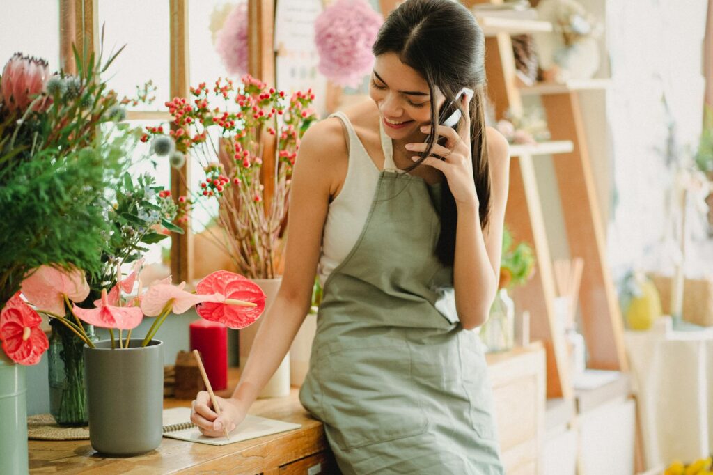 Female florist in apron on phone, writing notes in a floral shop, surrounded by colorful flowers.